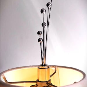 May include: A lamp finial featuring five thin, vertical silver-toned metal rods of varying heights, each topped with a small polished sphere. The finial is mounted on a cylindrical gold-toned base that sits atop a lampshade frame.