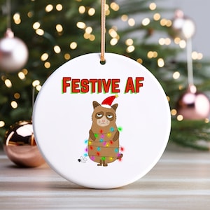 Könnte beinhalten: Ein weißer Keramik-Weihnachtsbaumschmuck mit einer Comic-Katze, die einen Weihnachtsmann-Hut trägt und in Weihnachtslichter gewickelt ist. Der Text "FESTIVE AF" ist in Rot und Grün auf dem Ornament gedruckt.