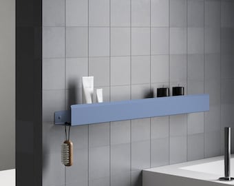 May include: A long, matte blue metal wall-mounted shelf featuring a raised front edge. The shelf holds two white tubes and two black cylindrical containers. A wooden-handled brush hangs from a small hook on the left side of the shelf.