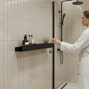 Puede incluir: Un estante de pared negro mate instalado sobre azulejos verticales de color claro. El estante sostiene una botella marrón, dos cepillos de dientes de madera, un tubo blanco pequeño y una pequeña planta suculenta verde en una maceta. Una ducha negra está cerca.