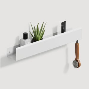 May include: A long, white, rectangular metal wall shelf with a front lip. The shelf holds a small square bottle with a black cap, a green succulent plant in a black pot, and a black tube. A wooden brush hangs from the right side of the shelf.