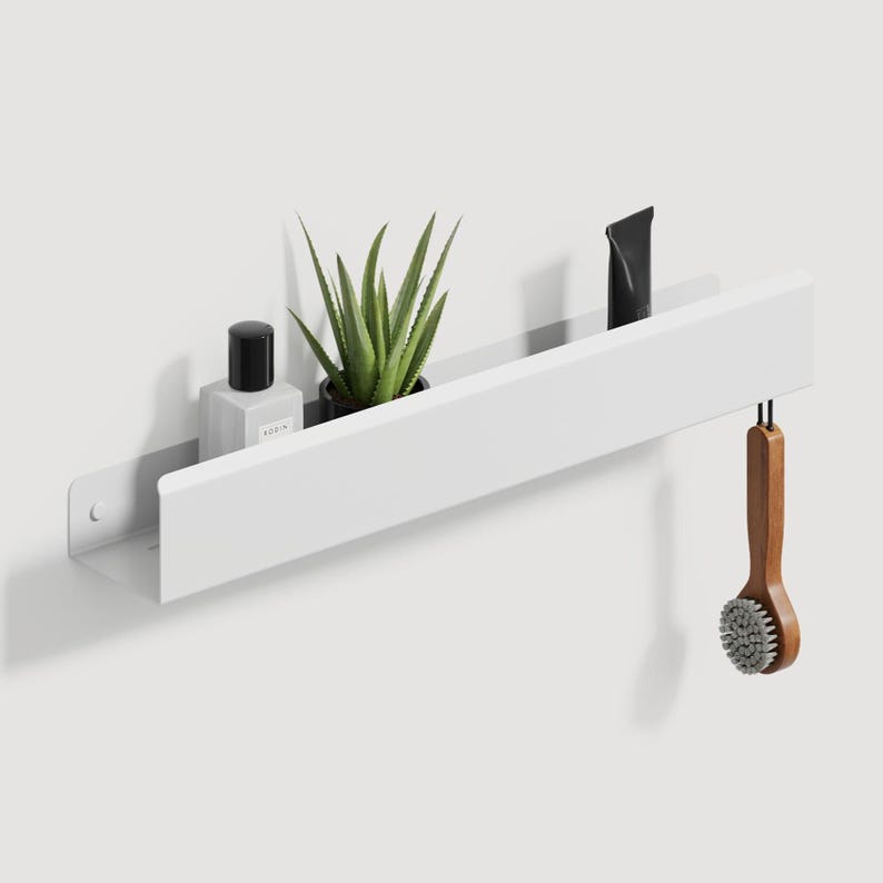 May include: A long, white, rectangular metal wall shelf with a front lip. The shelf holds a small square bottle with a black cap, a green succulent plant in a black pot, and a black tube. A wooden brush hangs from the right side of the shelf.
