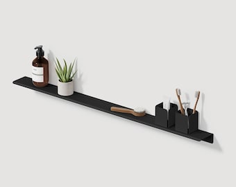 Può includere: Una mensola galleggiante in metallo nero con accessori per il bagno. La mensola contiene un dosatore di sapone marrone, una piccola pianta in vaso, una spazzola di legno e due contenitori neri con spazzolini e dentifricio.