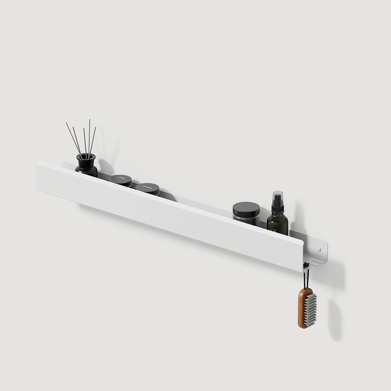 May include: A long, white, wall-mounted metal shelf with a flat front panel. The shelf holds a black reed diffuser, two small black circular containers, and a dark glass spray bottle. A small wooden brush hangs from a hook on the right side.