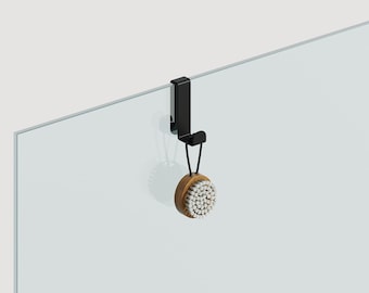May include: A black shower hook with a round, wooden bath brush hanging from it. The hook is attached to a glass shower door.