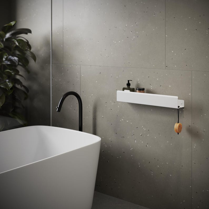 May include: A white rectangular wall-mounted shelf featuring a flat surface for storage and a small hook on the right side. A round tan soap bar hangs from the hook by a black cord. The shelf holds a dark pump bottle and a small jar.