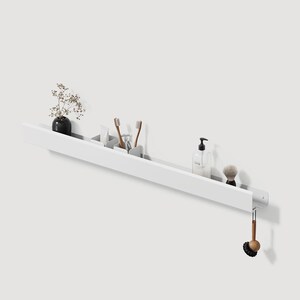 May include: A long, white, wall-mounted shelf featuring a slim profile. The shelf holds a small black vase with dried flowers, two small white containers with toothbrushes, a clear soap dispenser, a shaving brush, and a hanging wooden scrub brush.