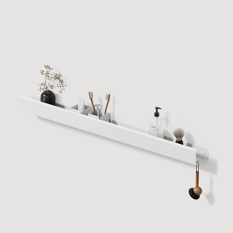 May include: A long, white, wall-mounted shelf featuring a slim profile. The shelf holds a small black vase with dried flowers, two small white containers with toothbrushes, a clear soap dispenser, a shaving brush, and a hanging wooden scrub brush.
