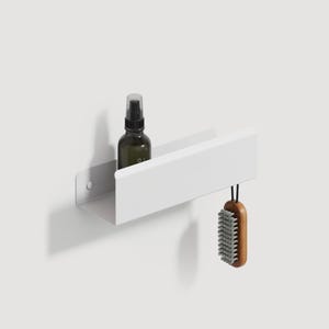 May include: A white wall-mounted metal shelf with a rectangular profile. A dark amber glass spray bottle sits inside the shelf, and a wooden-handled brush with grey bristles hangs from the right side of the shelf by a black cord.
