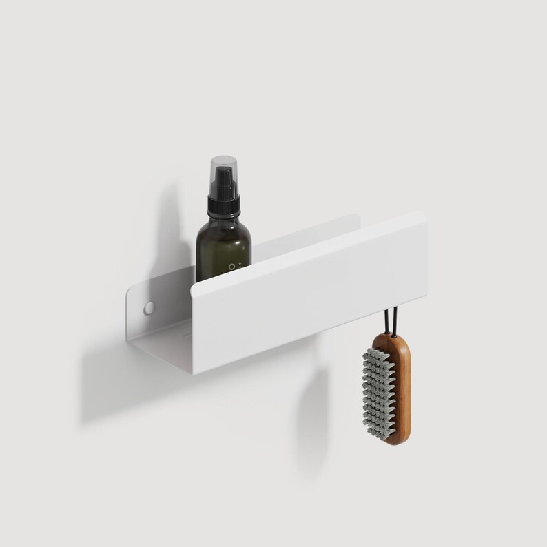 May include: A white wall-mounted metal shelf with a rectangular profile. A dark amber glass spray bottle sits inside the shelf, and a wooden-handled brush with grey bristles hangs from the right side of the shelf by a black cord.