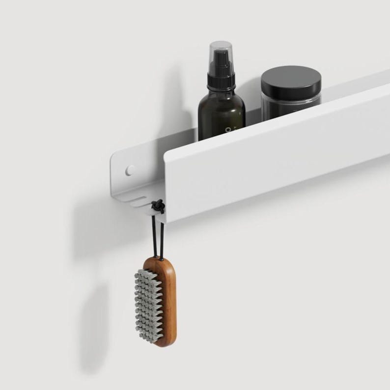 May include: A white wall-mounted metal shelf with a long horizontal tray. The shelf holds a dark amber spray bottle and a black jar. A wooden nail brush with grey bristles hangs from a small black cord attached to the underside of the shelf.