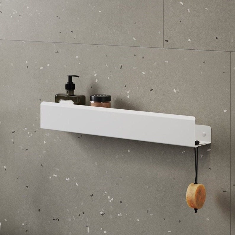 May include: A white wall-mounted shelf with a rectangular profile and a flat top surface. The shelf holds a dark pump bottle and a small jar. A round, light-coloured object hangs from a black cord attached to the right side of the shelf.