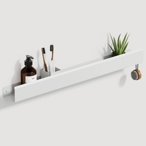 May include: A long, white, wall-mounted metal shelf holding a brown amber glass pump bottle, a white square container with two toothbrushes and a tube, a small green succulent plant in a black pot, and a round wooden brush hanging from the side.