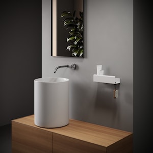 May include: A white cylindrical vessel sink sits on a light wood vanity. Above, a wall-mounted faucet and a rectangular mirror reflect a green plant. A white wall-mounted shelf holds two small white tubes, with a wooden brush hanging from a hook.