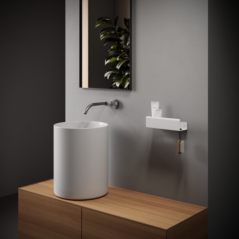May include: A white cylindrical vessel sink sits on a light wood vanity. Above, a wall-mounted faucet and a rectangular mirror reflect a green plant. A white wall-mounted shelf holds two small white tubes, with a wooden brush hanging from a hook.