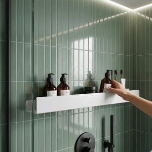 May include: A white, rectangular, wall-mounted shower shelf with a speckled finish. The shelf holds two amber-colored bottles with white labels, a small black jar, a brown bottle, and two toothbrushes. It is mounted against sage green tiles.