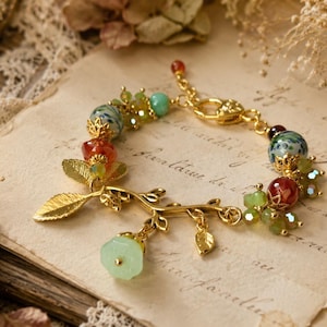 Könnte beinhalten: Ein goldfarbenes Bettelarmband mit einer zweigartigen Struktur, metallischen Blättern und einem hellgrünen Blumenanhänger. Das Armband ist mit mehrfarbigen Perlen in Rot-, Grün- und Blautönen verziert und verfügt über einen Karabinerverschluss.