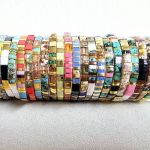 Peut inclure: Une collection de bracelets de perles colorées. Chaque bracelet est composé de petites perles rectangulaires de différentes couleurs, dont le turquoise, le noir, le blanc, le rouge et l'or. Les bracelets sont empilés, présentant une variété de motifs.