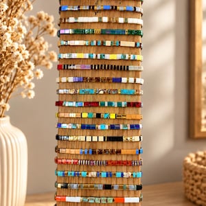 Può includere: Un espositore verticale in legno che sostiene una collezione di ventidue braccialetti impilati. Ogni braccialetto è composto da piccole perline quadrate multicolori con vari motivi, inclusi colori solidi, finiture metalliche e disegni marmorizzati.