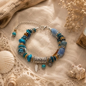 Peut inclure: Un bracelet multi-brins composé de perles turquoise, bleues et brunes, incluant des formes florales sculptées et une perle tubulaire centrale en métal argenté. Le bijou comporte de petites breloques en forme de plume et de fleur, avec un fermoir.