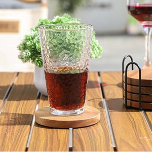 May include: A glass of dark brown liquid on a wooden coaster. The glass has a textured surface and is sitting on a wooden table. There is a set of wooden coasters in a black metal holder to the right of the glass.