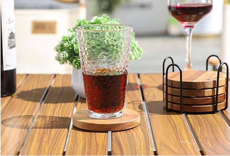 May include: A glass of dark brown liquid on a wooden coaster. The glass has a textured surface and is sitting on a wooden table. There is a set of wooden coasters in a black metal holder to the right of the glass.