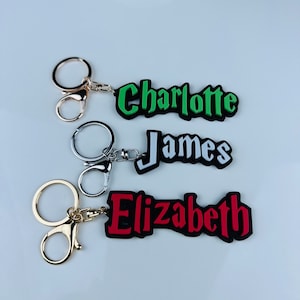 May include: Three personalized keychains with names: "Charlotte" in green, "James" in white, and "Elizabeth" in red. Each name is on a black background with a silver or gold-colored keyring and clasp.