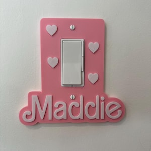 May include: A pink light switch cover with white hearts and the name "Maddie" in white script. The switch cover is mounted on a white wall. The switch itself is white.