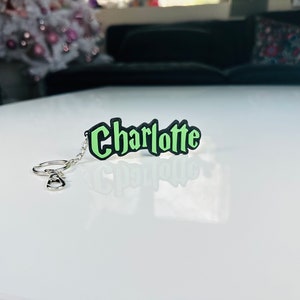 May include: A green and black keychain with the name "Charlotte" in a stylized font. The keychain has a silver-colored chain and ring. The name is in a bold, outlined font.