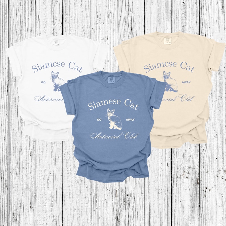 以下が含まれることがあります： 「Siamese Cat Go Away Antisocial Club」というテキストと、シャム猫のグラフィックがプリントされたTシャツが3枚。Tシャツの色は白、ベージュ、青です。