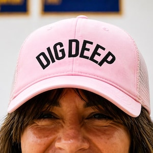 Op de afbeelding: Een lichtroze truckerpet met de woorden "DIG DEEP" in vette zwarte letters over de voorkant. De pet heeft een mesh achterkant en een gebogen klep. De pet wordt door een persoon gedragen.