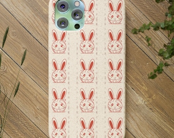 以下が含まれることがあります： ベージュ色のスマートフォンケースで、斑点のある質感に、長い耳と小さな星のアクセントが付いた赤い線画のウサギの顔が繰り返されるパターンが特徴です。ケースの左上隅には、3つのレンズを備えたカメラシステム用の切り欠きがあります。