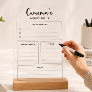 Może przedstawiać: Tygodniowa tablica planowania z przezroczystego akrylu z drewnianą podstawą. Tablica zawiera nadrukowany czarny tekst, w tym Cameron's Weekly Focus, Top 3 Priorities, Appointments, To Do i Notes, z polami wyboru i liniami do zapisywania zadań.