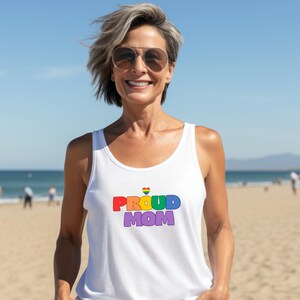 Op de afbeelding: Witte tanktop met de tekst "PROUD MOM" in regenboogkleuren en een regenbooghart. De tanktop wordt gedragen door een persoon op een zonnige stranddag.