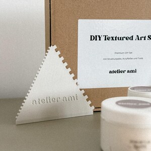 May include: A DIY textured art set with a white triangular tool, a brown box, and white containers. The text "DIY Textured Art Set" and "atelier ami" are visible on the box and tool. The set includes premium DIY supplies.