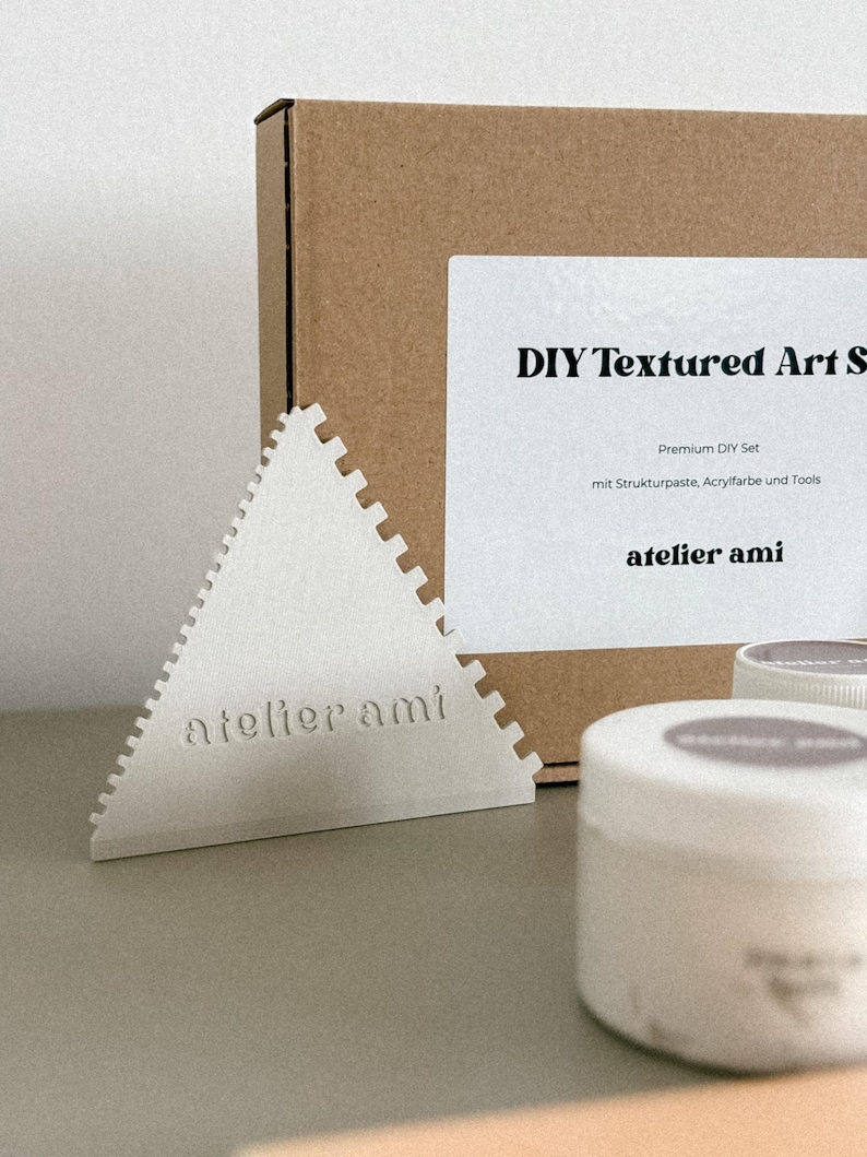 May include: A DIY textured art set with a white triangular tool, a brown box, and white containers. The text "DIY Textured Art Set" and "atelier ami" are visible on the box and tool. The set includes premium DIY supplies.