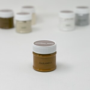 May include: A small, clear jar of caramel-colored paint with a white lid. The jar is labeled "KARAMELL" in a dark brown font. The brand name "atelier ami" is printed on the lid. Several other jars of paint are visible in the background.