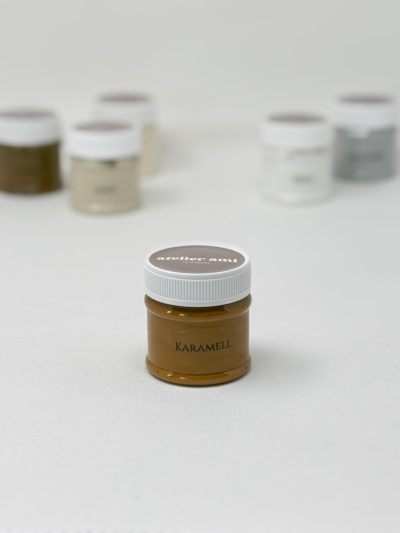 May include: A small, clear jar of caramel-colored paint with a white lid. The jar is labeled "KARAMELL" in a dark brown font. The brand name "atelier ami" is printed on the lid. Several other jars of paint are visible in the background.