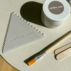 May include: A light gray triangular tool with a notched edge and the text "atelier ami" is next to a white container with a brown label. A paintbrush and two wooden sticks are also present. The items are on a light-colored surface.