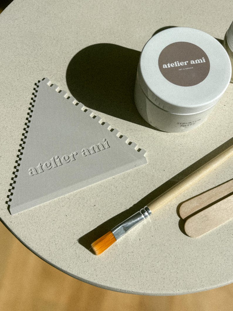 May include: A light gray triangular tool with a notched edge and the text "atelier ami" is next to a white container with a brown label. A paintbrush and two wooden sticks are also present. The items are on a light-colored surface.