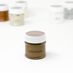 May include: A small, round jar of chocolate-colored paint with a white lid. The word "CHOCOLATE" is printed on the front of the jar. Other jars are visible in the background.