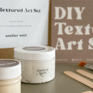 May include: A DIY textured art set with beige and white jars of paste, wooden stir sticks, and a paintbrush. The set includes a box with the words "DIY Textured Art Set" and a card that says "Textured Art Set".