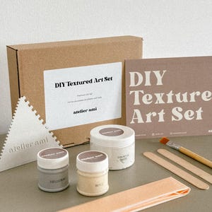 May include: A DIY Textured Art Set with various art supplies. The set includes a cardboard box, containers of paint, a triangular tool, a brush, and wooden sticks. The set is labeled "DIY Textured Art Set" and "atelier ami".