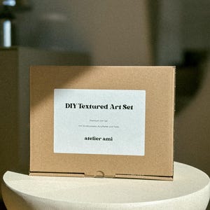 May include: A brown cardboard box labeled "DIY Textured Art Set" with white text. The box includes "Premium DIY Set" and "atelier ami" text. The box is sitting on a white, rounded pedestal.