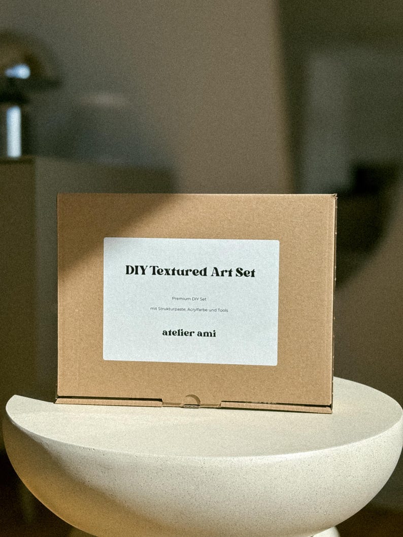 May include: A brown cardboard box labeled "DIY Textured Art Set" with white text. The box includes "Premium DIY Set" and "atelier ami" text. The box is sitting on a white, rounded pedestal.