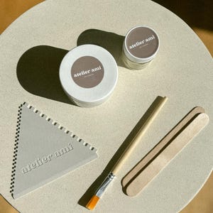 May include: A collection of art supplies, including two small jars with brown labels, a triangular tool, a paintbrush, and two wooden sticks. The jars and tool have the text "atelier ami". The items are arranged on a light-colored, round surface.