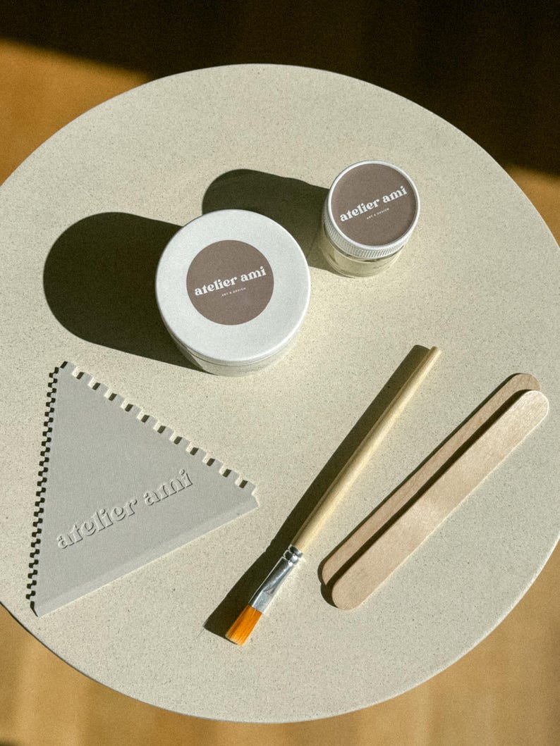 May include: A collection of art supplies, including two small jars with brown labels, a triangular tool, a paintbrush, and two wooden sticks. The jars and tool have the text "atelier ami". The items are arranged on a light-colored, round surface.