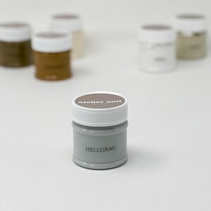 May include: A small, gray paint jar with a white lid, labeled "HELLGRAU." The jar is in focus, with other jars of paint in various neutral colors blurred in the background. The brand name "atelier ami" is printed on the lid.