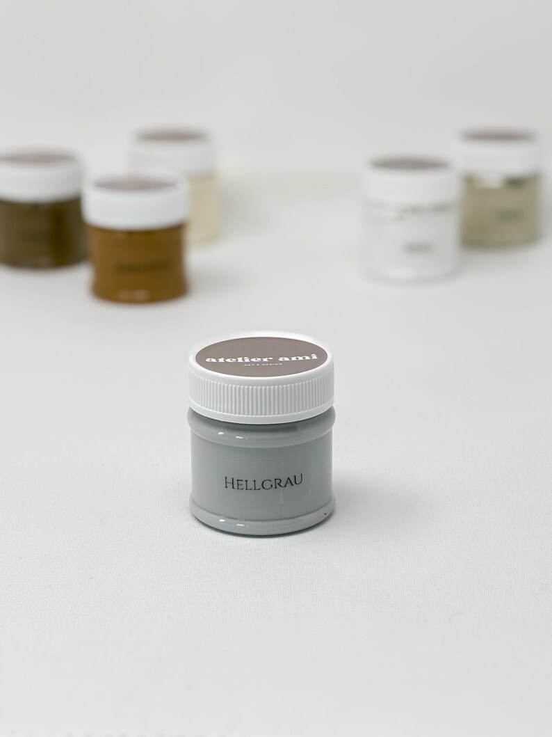 May include: A small, gray paint jar with a white lid, labeled "HELLGRAU." The jar is in focus, with other jars of paint in various neutral colors blurred in the background. The brand name "atelier ami" is printed on the lid.