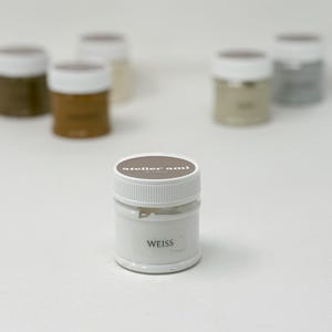 May include: A collection of small, white paint jars with white lids. The front jar has the word "WEISS" printed on it. The lids are brown and have the text "atelier ami" printed on them. Other jars in the background are various shades of brown and gray.